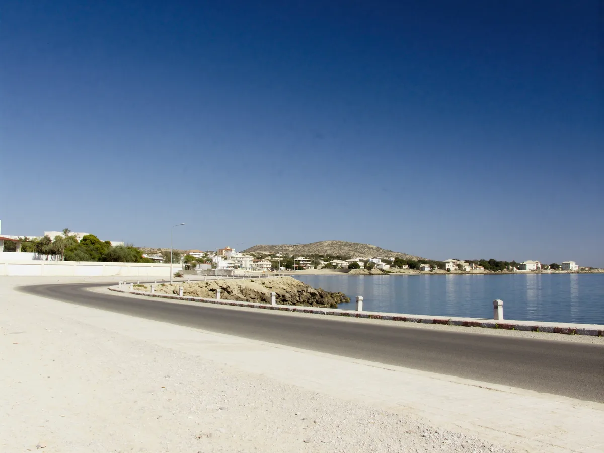 AI-generated image: a quiet stretch of rocky shoreline and low white houses near pilar de jaravía under clear winter light, empty road, no people