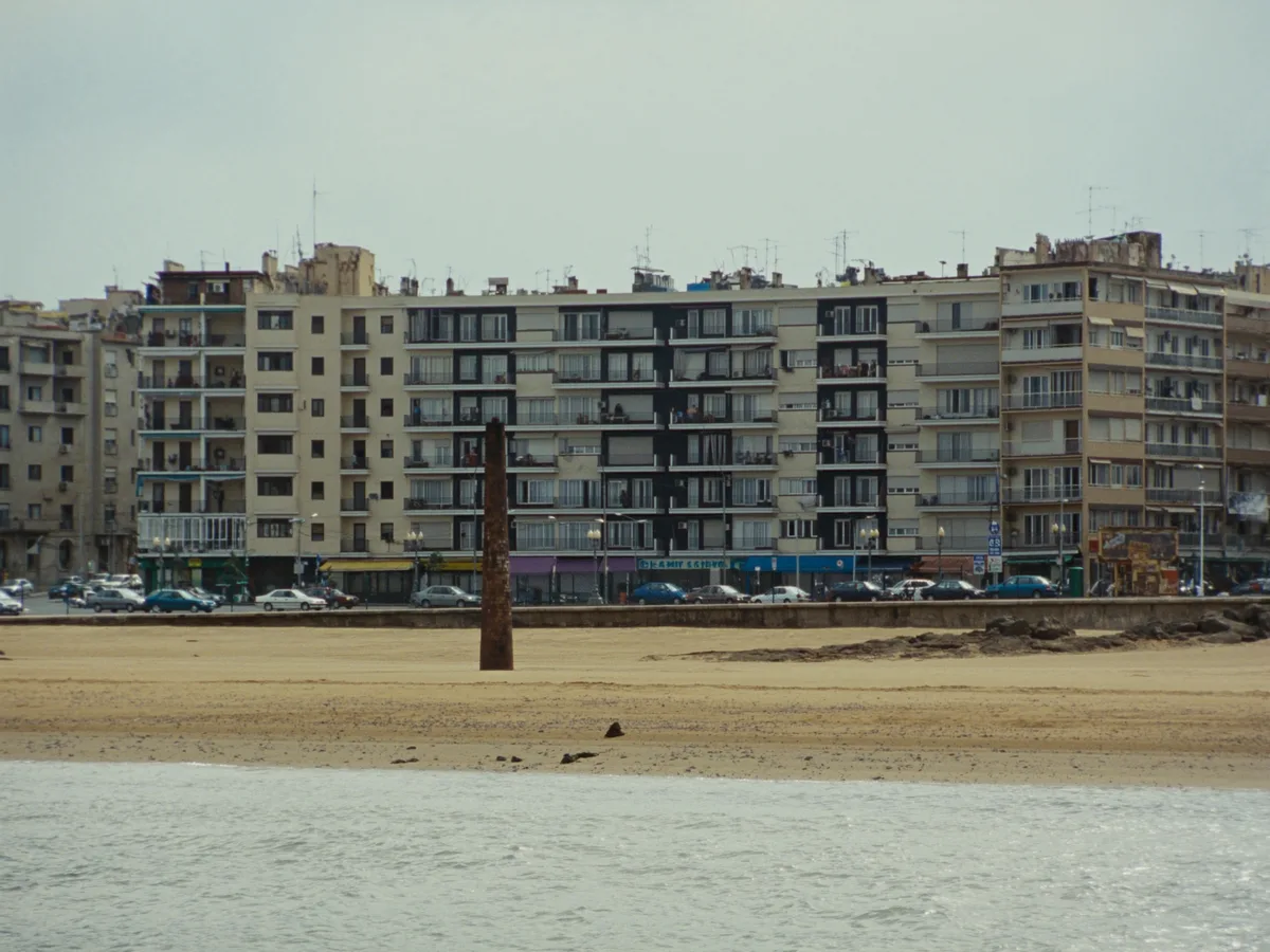 AI-generated image: a working seafront of mid-century apartment blocks behind a wide beach and a lone brick chimney, flat grey-blue sea, soft overcast light