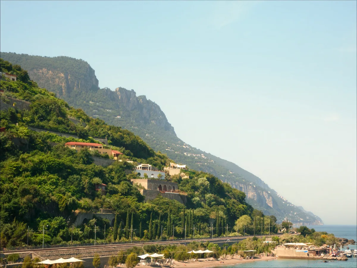 AI-generated image: terraced vineyards and villas on a green hillside dropping towards the mediterranean, a rail line and a narrow beach below, clear morning light