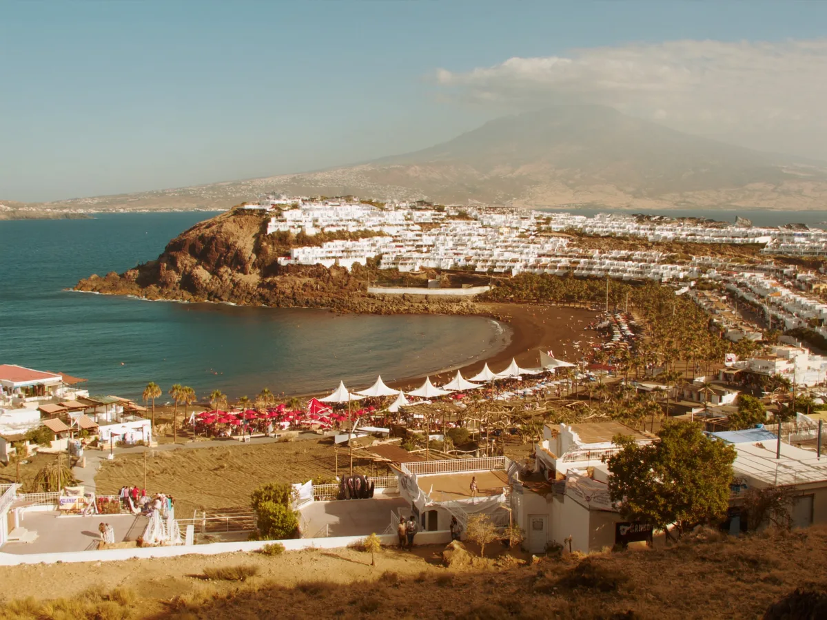 AI-generated image: sunlit low-rise whitewashed apartments above a dark volcanic-sand beach, typical of the Costa Adeje coast in Tenerife