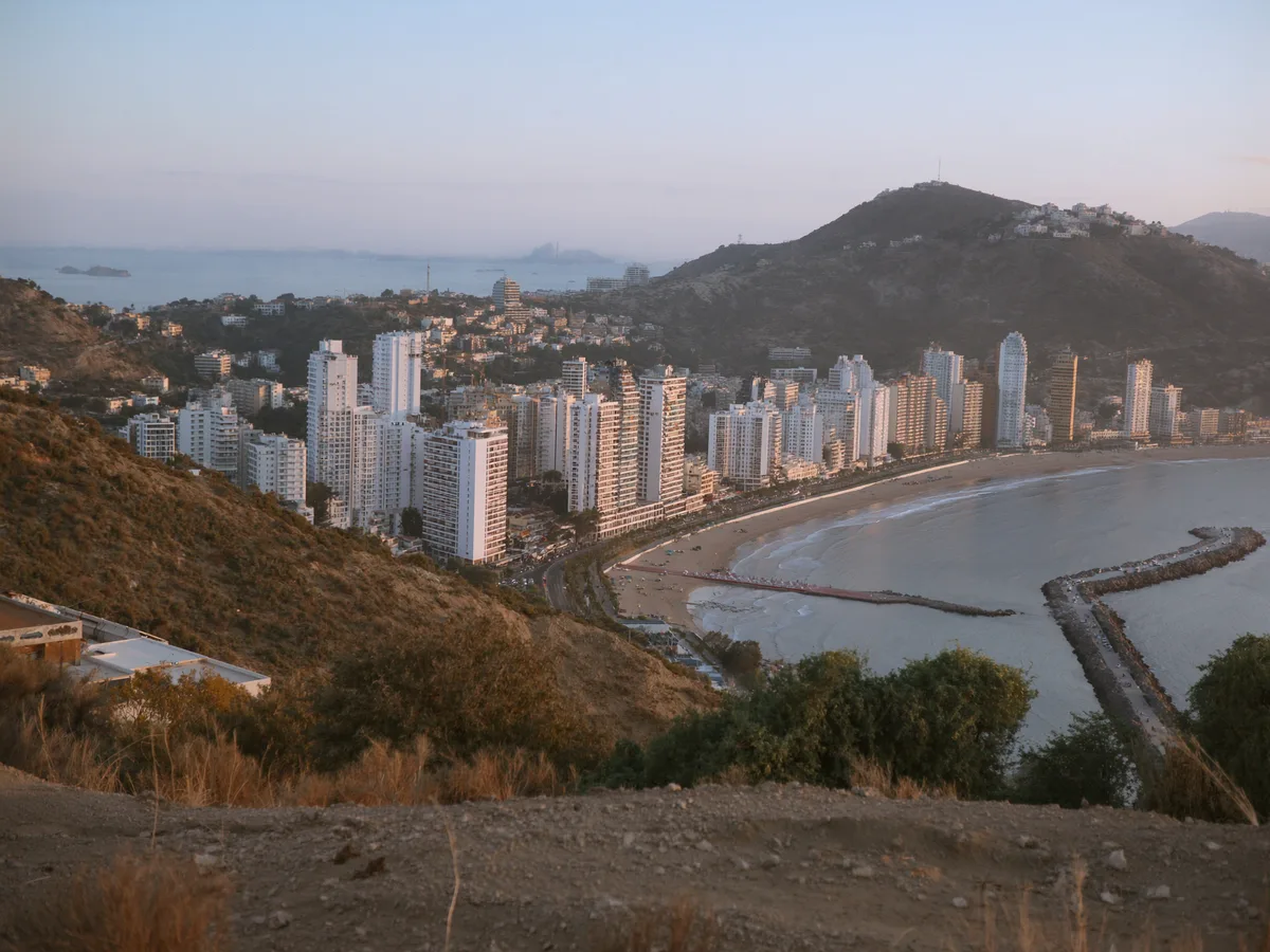 AI-generated image: a dense cluster of tall white towers along a curved bay seen from a hillside at dusk, mountains behind, no people, no logos