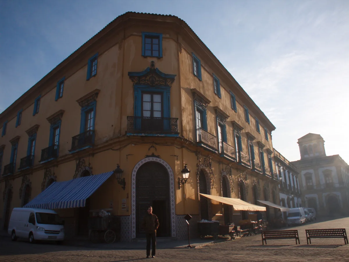 AI-generated image: a narrow street of tall tiled valencian facades with wrought-iron balconies opening onto a small plaza, soft morning light, no people, no logos