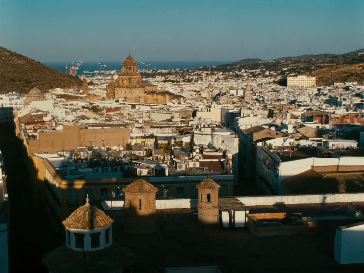 AI-generated image: a dense grid of city rooftops with a cathedral tower and a working port beyond, hills at the edge of frame, warm light, no people, no logos