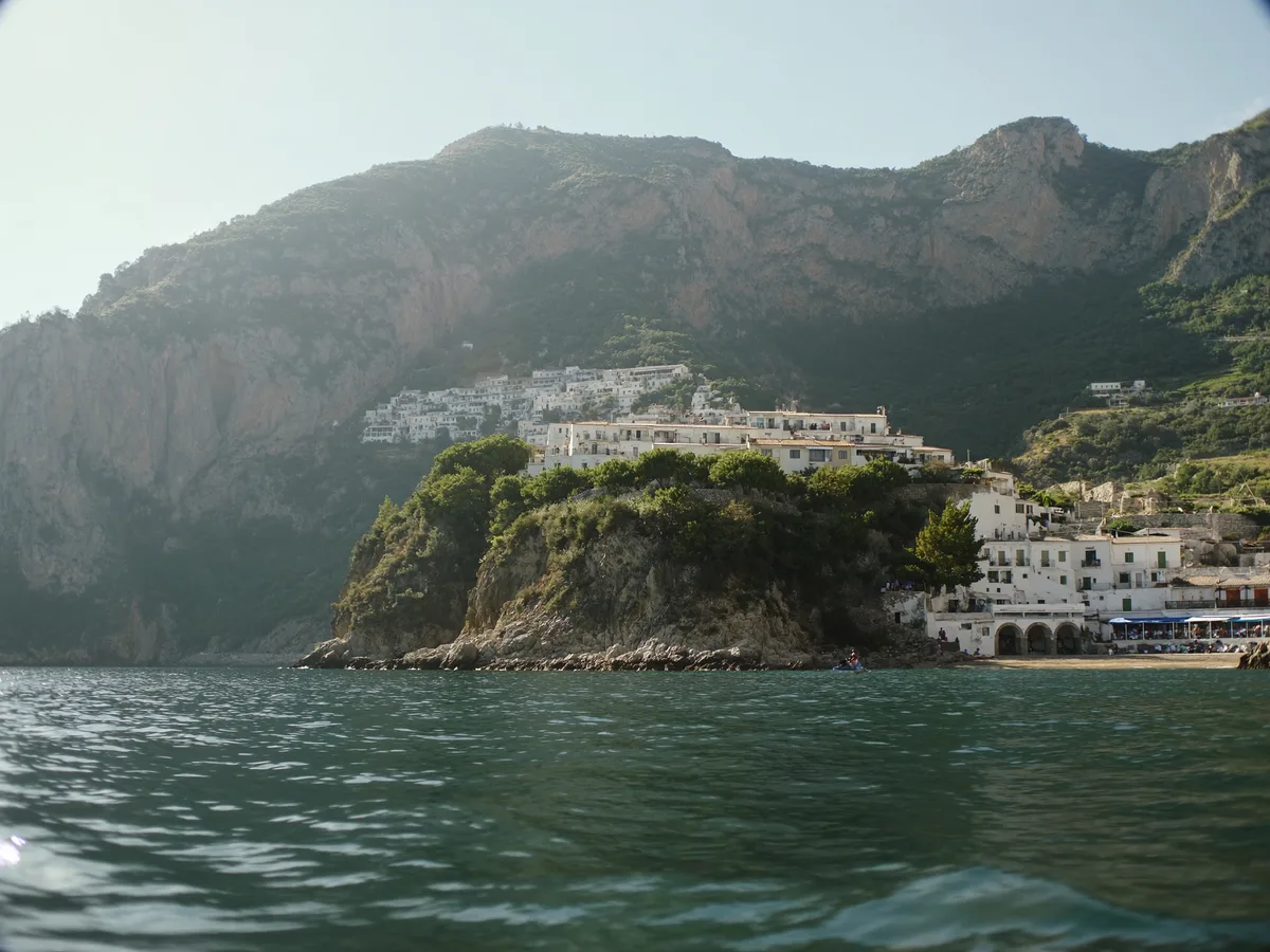 AI-generated image: a whitewashed clifftop town above a small cove with mountains rising directly behind, late afternoon light, no people, no logos