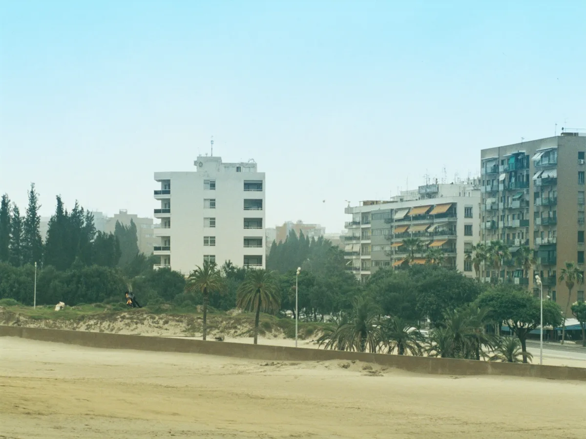 AI-gegenereerde afbeelding: een breed vlak strand met appartementen uit de jaren zestig achter duinen en lage pijnbomen, heiig blauw licht, geen mensen, geen logo&rsquo;s