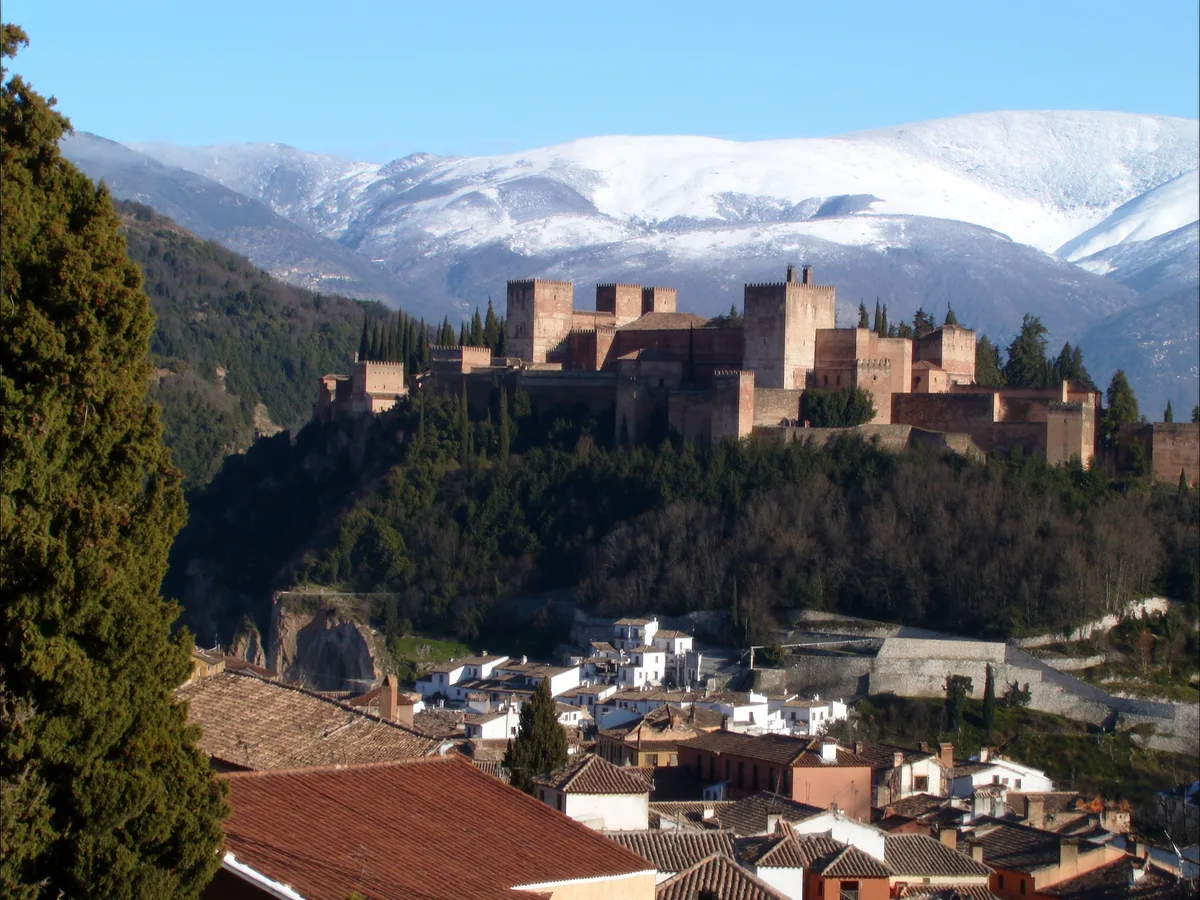 AI-gegenereerde afbeelding: het Alhambra met de sneeuwgrens van de Sierra Nevada erachter, gezien over de pannendaken van Granada in helder winterlicht, geen mensen