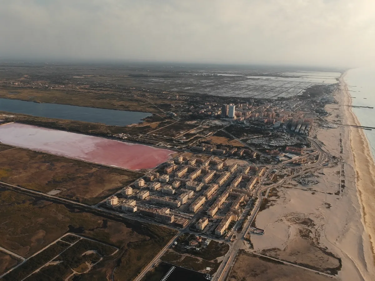KI-generiertes Bild: Luftaufnahme eines rosafarbenen Salzsees neben niedrigen wei&szlig;en Apartmenth&auml;usern und einem langen Sandstrand, flaches Land, sp&auml;tes Nachmittagslicht, keine Menschen, keine Logos