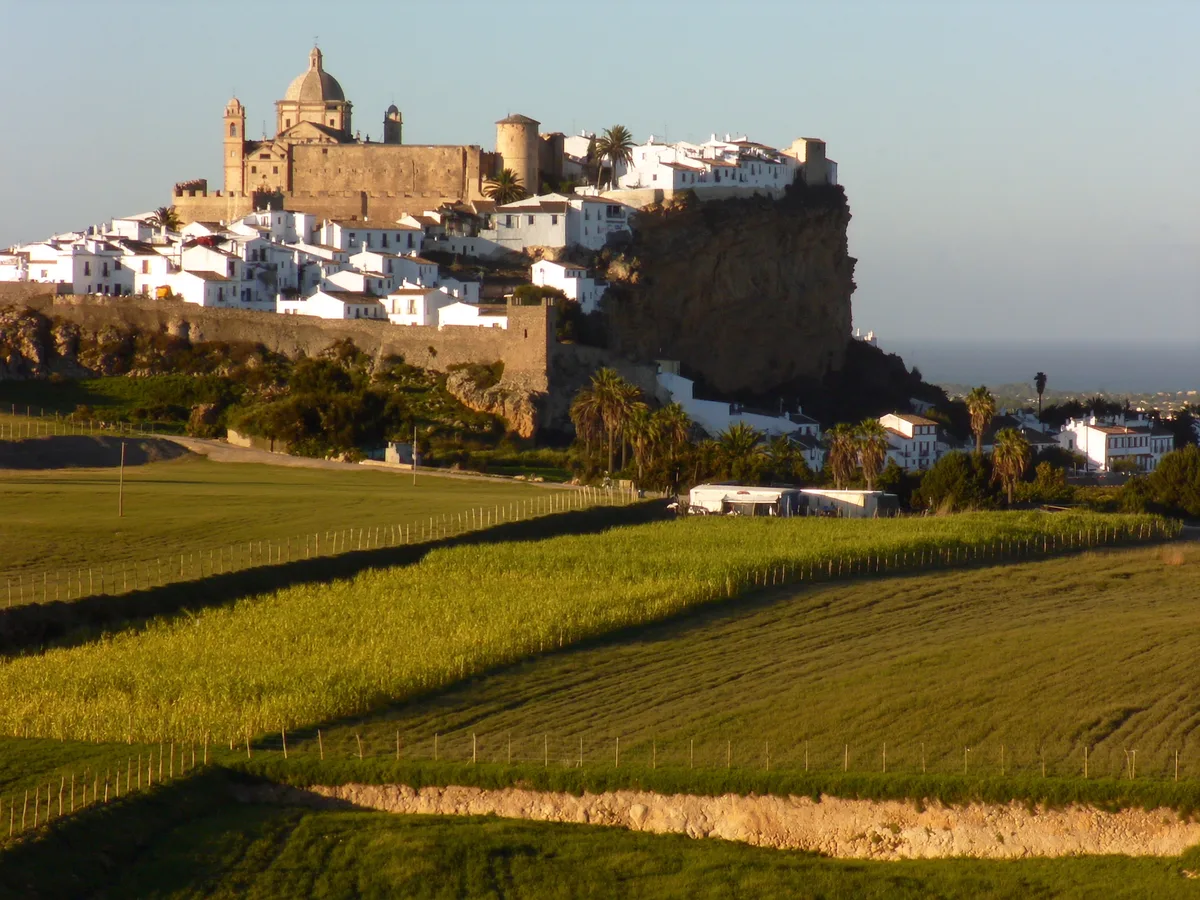 KI-generiertes Bild: die weißen Häuser und die Burg von Salobreña auf ihrem Felsen über grünen Zuckerrohrfeldern und dem Meer, warmes spätes Licht, keine Menschen