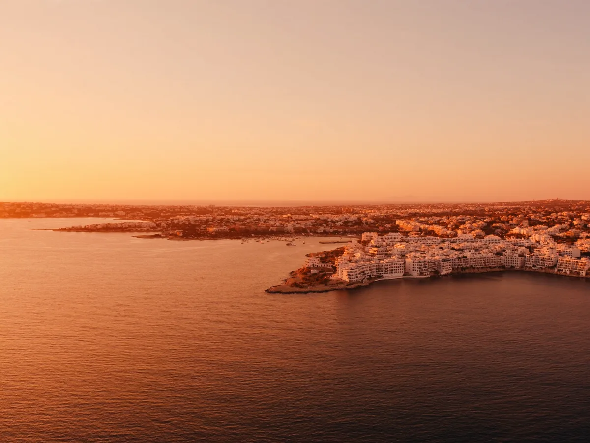 KI-generiertes Bild: eine weite, nach Westen offene Bucht bei Sonnenuntergang, niedrige weiße Apartmentterrassen im Bogen entlang der Küste und glattes orangefarbenes Wasser, keine Menschenmengen oder Schilder