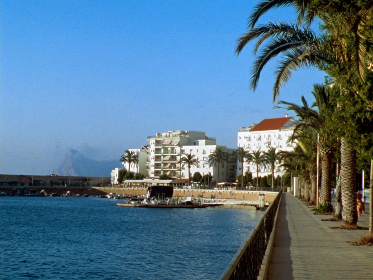 KI-generiertes Bild: ein weiter Morgenblick entlang einer palmengesäumten Uferpromenade mit einem kleinen Jachthafen und niedrigen weiß getünchten Apartmenthäusern, spiegelglattes Wasser, keine Menschen