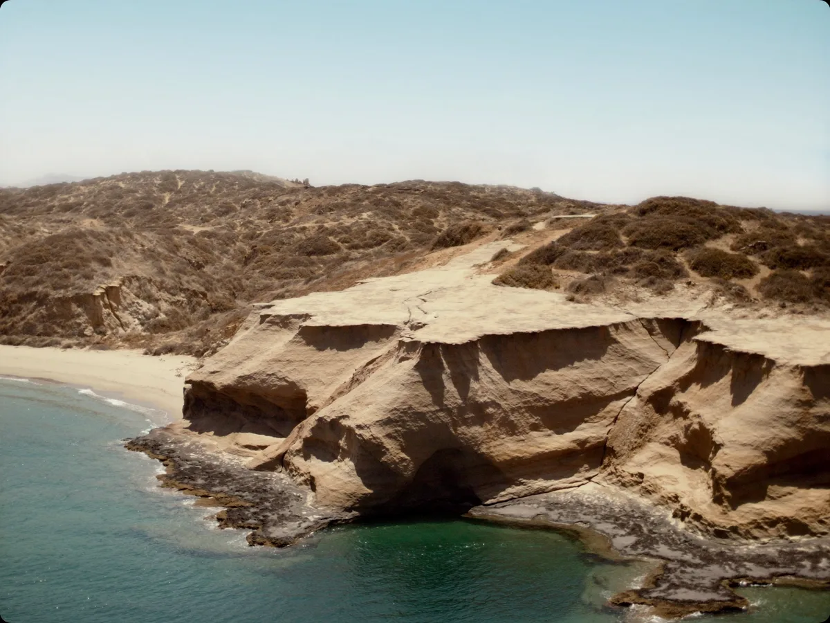 KI-generiertes Bild: eine felsige Landspitze, die neben einer leeren Bucht mit hellem Sand in klares Wasser abfällt, dahinter trockene, buschbewachsene Hügel, hartes Nachmittagslicht, keine Menschen, keine Logos