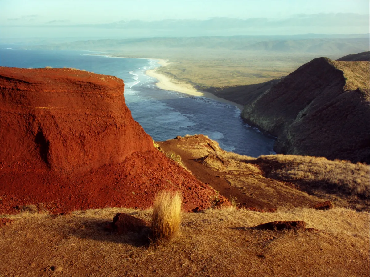 AI-generated image: red iron-rich cliffs above a narrow empty cove in early morning light, juniper scrub on the headland and a rough coastal track